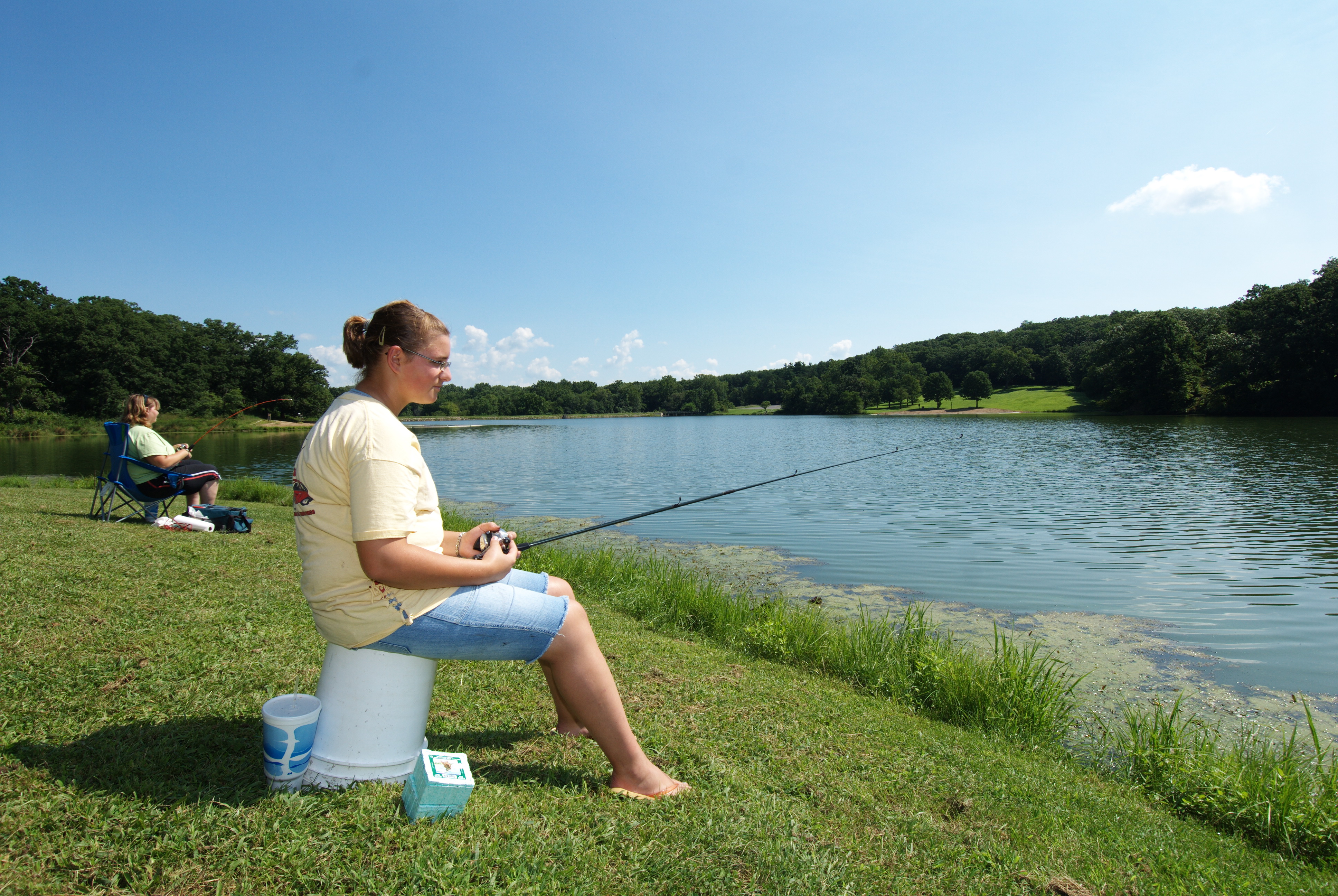 a lady sitting on a bucket along the shore of the lake fishing