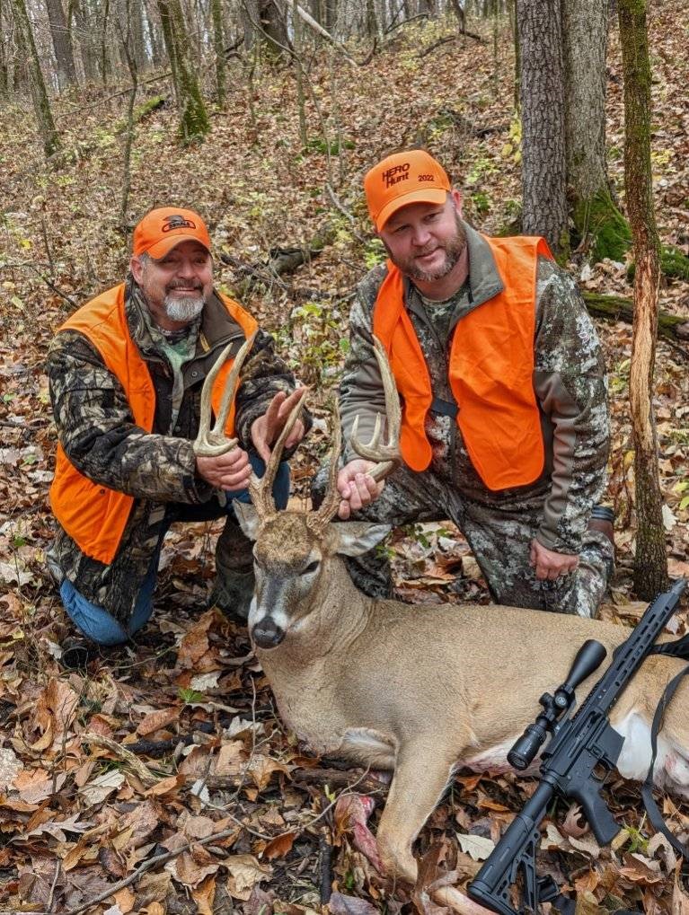 A veteran and a guide in camouflage and hunter orange pose in the woods with a large buck, the recipient of the Biggest Buck Award for 2021.