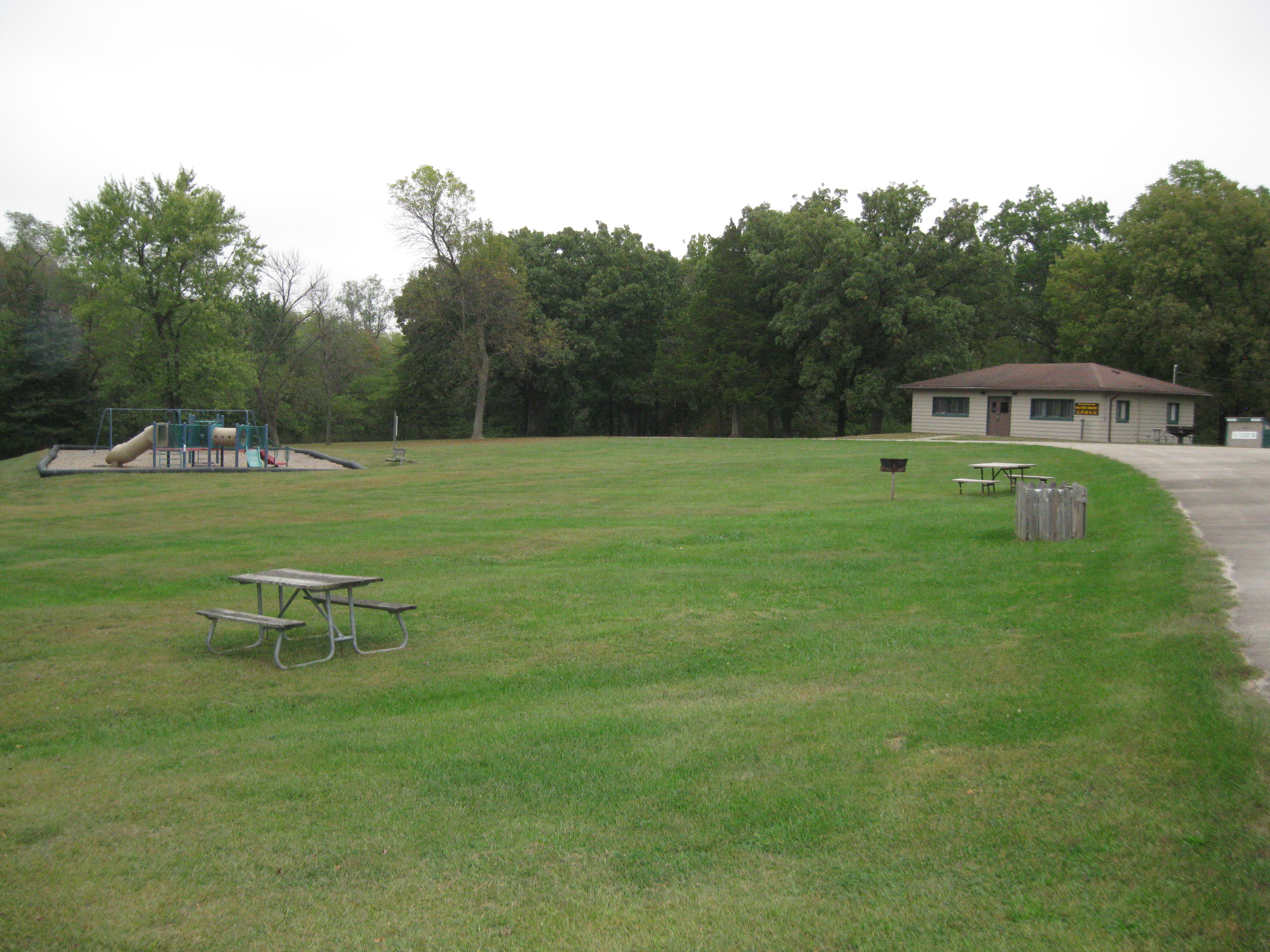 Picnic tables, playground and grill in field and enclosed shelter in the distance