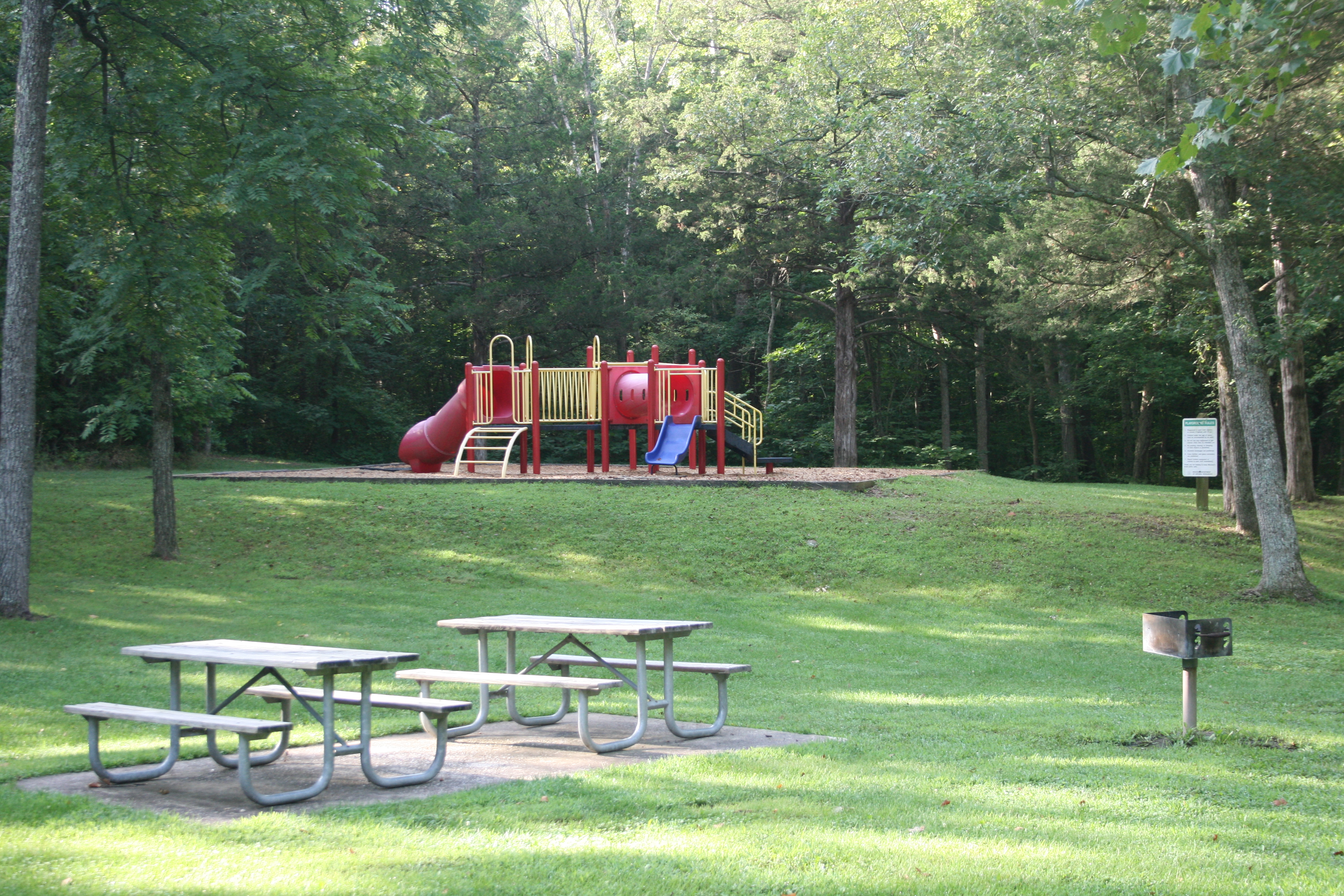 two picnic tables on a concrete pad and a grill with a playground in the background