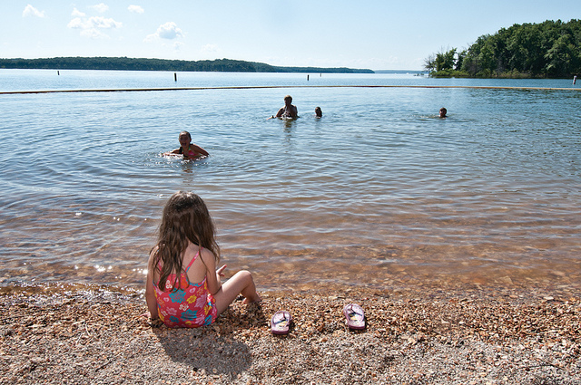 a little girl sits on the beach watching other kids swim