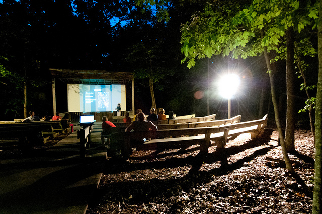 people sit in the dark on the wooden benches of the amphitheater watching a program