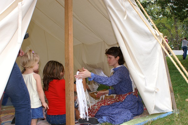 lady in period costume showing kids clothing items inside a tent