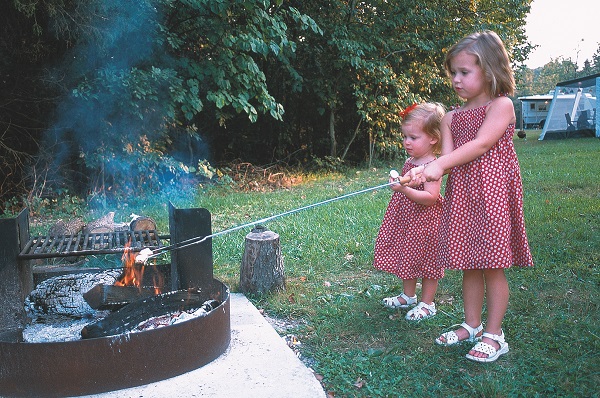 two toddler girls in matching dresses roasting marshmallows over a campfire