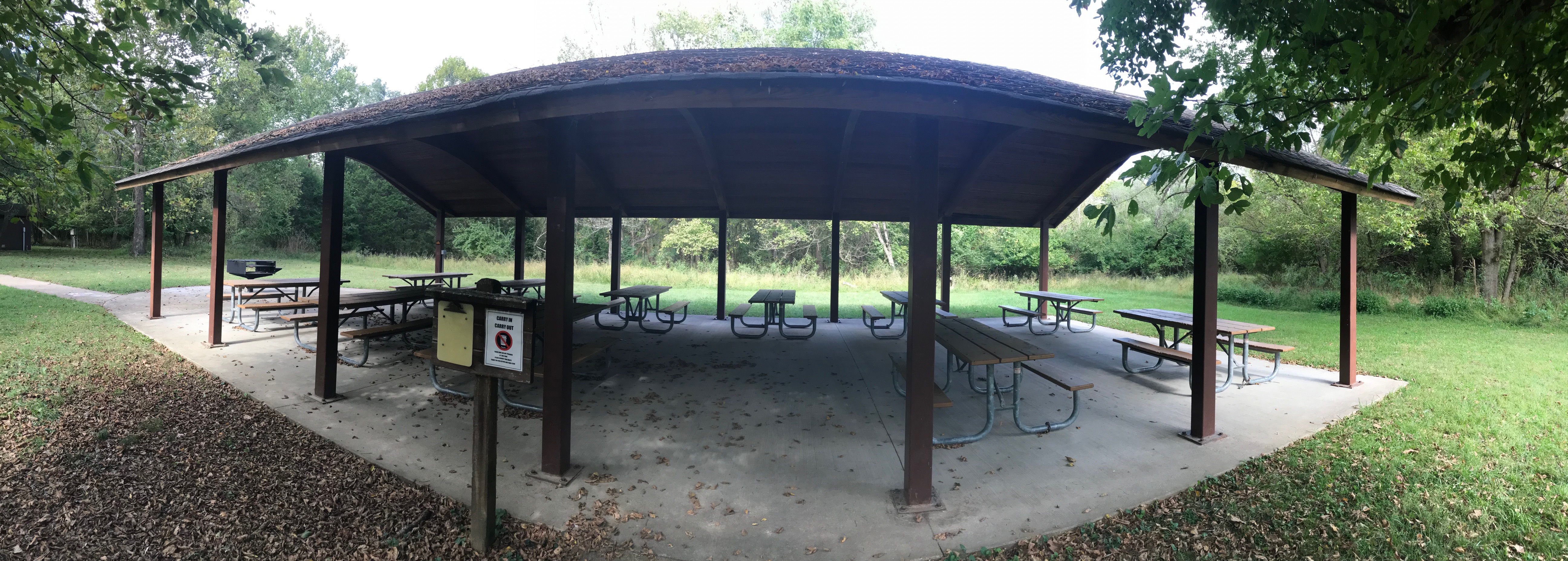 Panoramic view of an open shelter, with several picnic tables under the shelter roof and a grill to the left