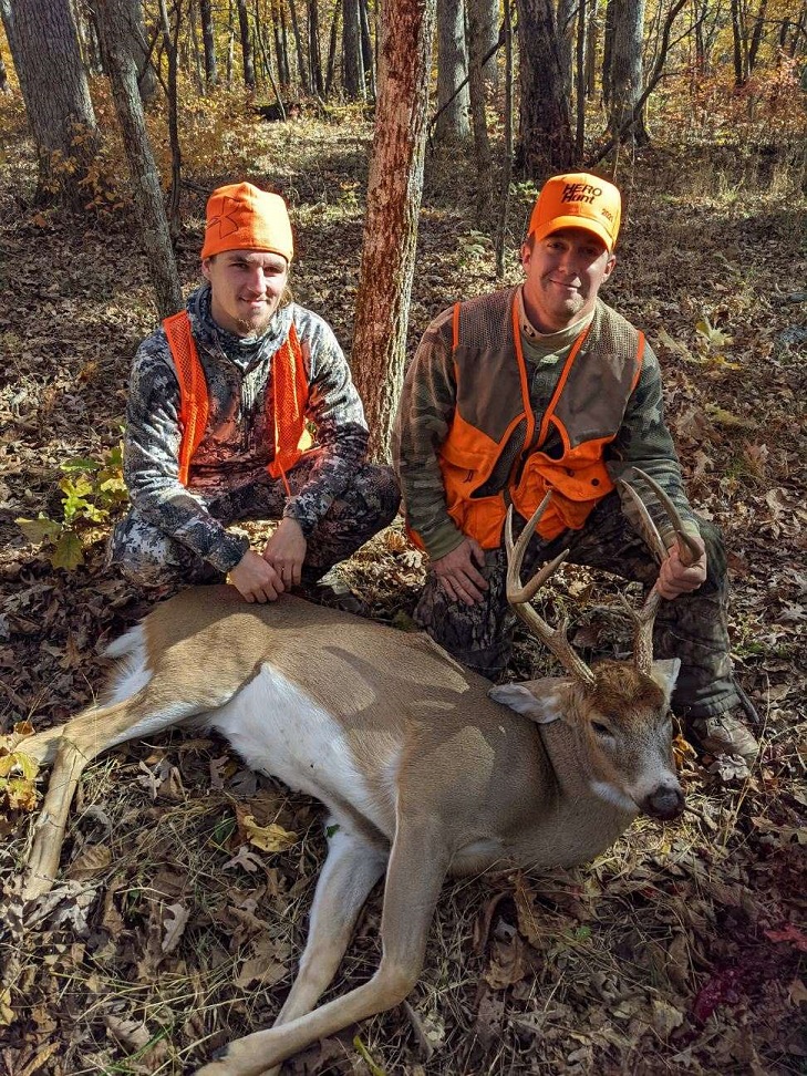 A veteran and a guide, both in camouflage and hunter orange pose with a harvested buck in the woods. 