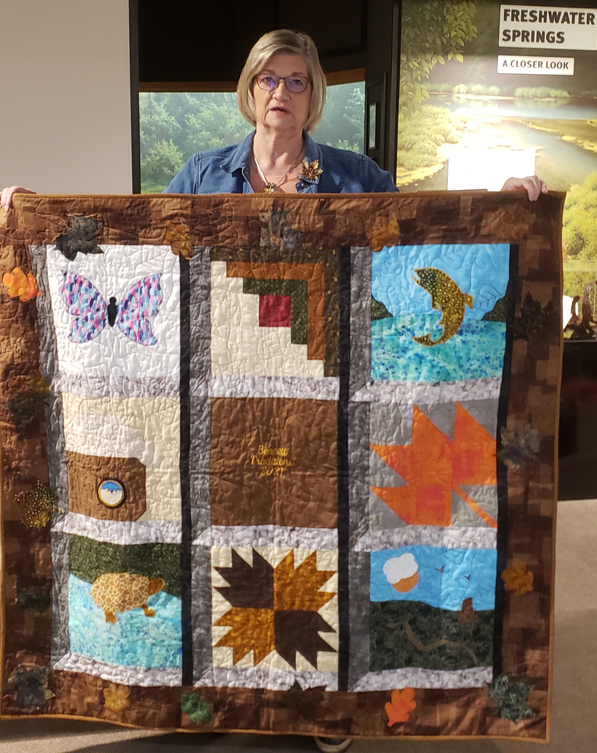 A woman holds up a quilt with designs including a butterfly, a trout, a leaf and a beaver