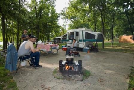 a couple cooking over their campfire with their picnic table and camper in the background