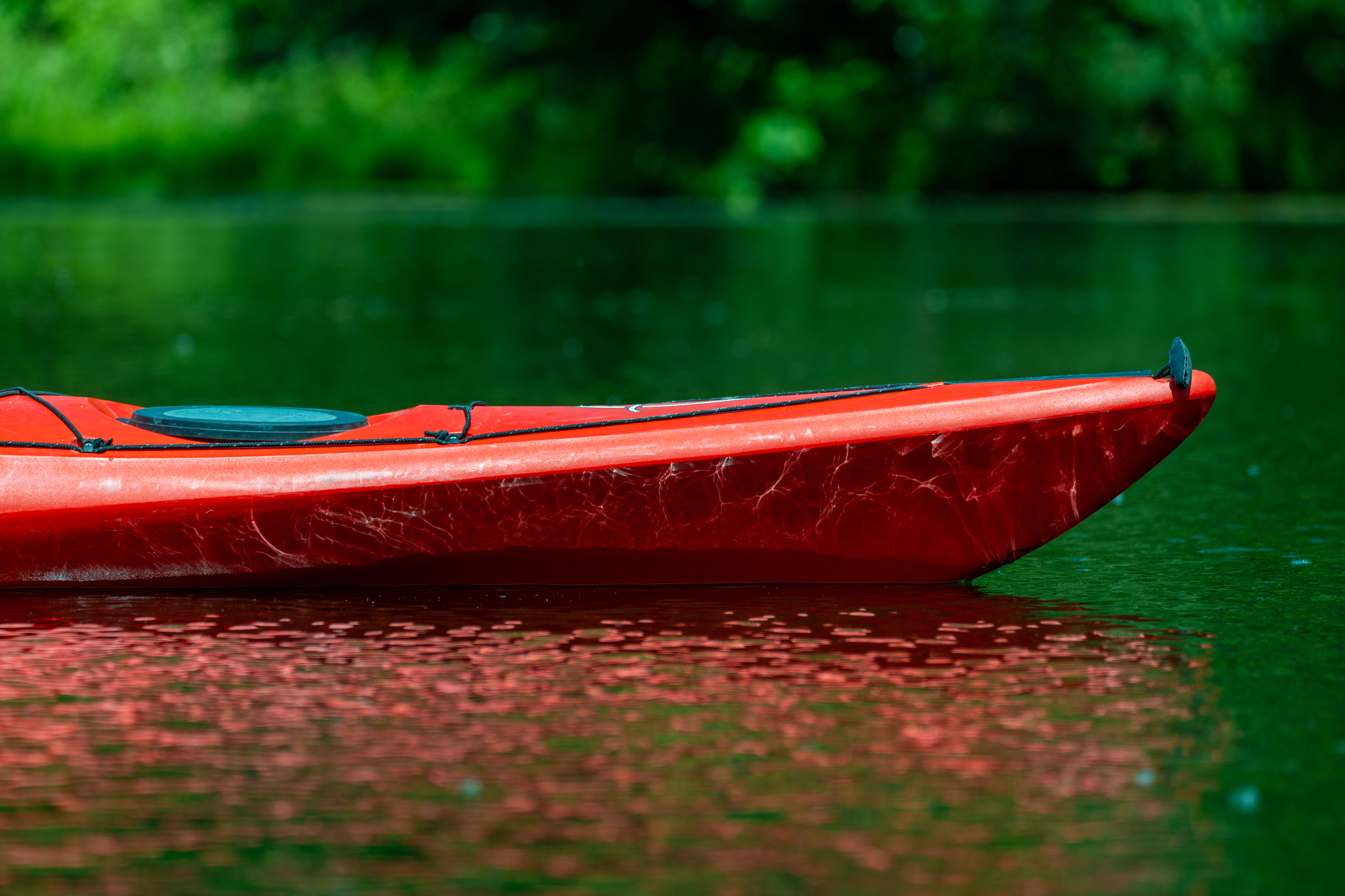 A close-up image of a reddish-orange kayak floating on a calm body of water with trees in the background